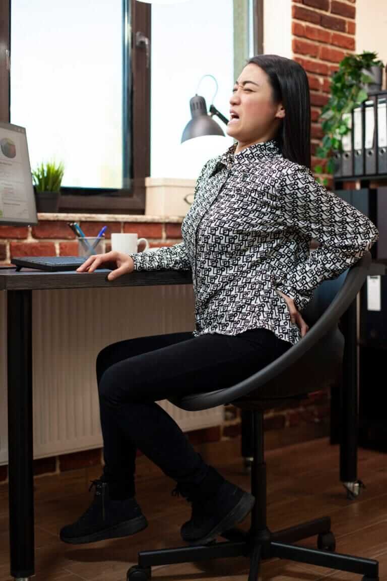 Asian lady grabs her back in pain while sitting on an office chair